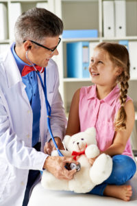 Joyful girl looking at her doctor examining teddy bear in hospital